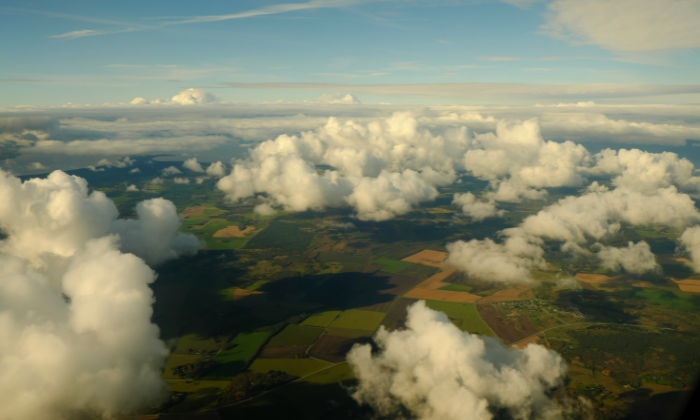 Aerial view of green farmland and forests partly covered by fluffy white clouds under a blue sky.