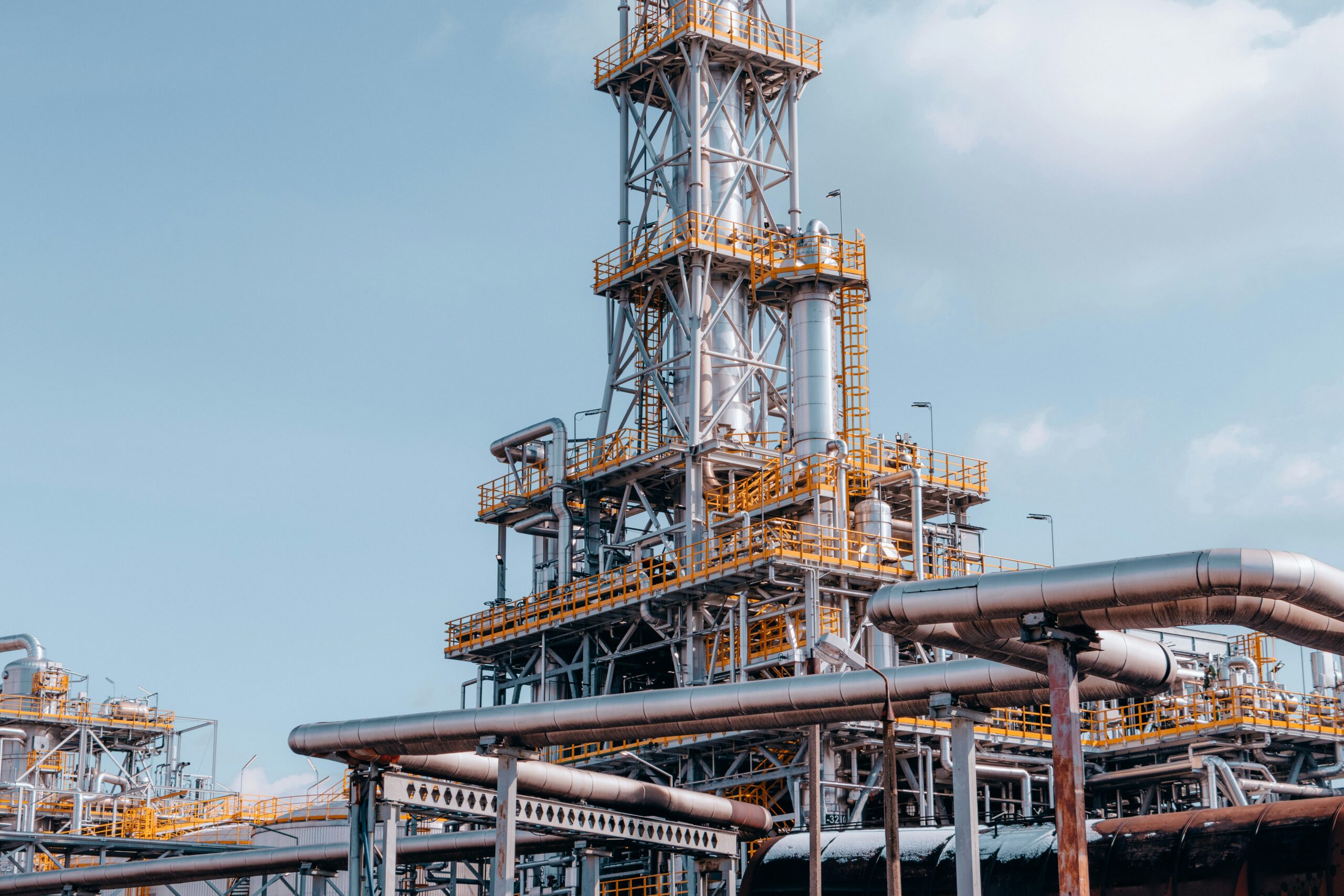 An industrial processing facility against a pale blue sky, with tall stainless-steel distillation columns, bright yellow access platforms and safety railings, and networks of large insulated pipes running across the foreground. The Investment Booster finances industrial decarbonisation, which is why an industrial facility is shown.